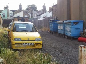 Yellow car parked beside blue commercial bins behind a row of South London houses