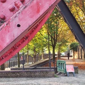 A riverside path in South London framed by the red and black beams of a bridge, with a lone figure walking beneath autumn trees.